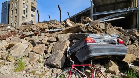 SARAID The rear end of a silver car and a damaged red bike sticking out from underneath a mountain of concrete rubble. In the background there is a tall concrete building and the metal skeleton of a multi storey car park.