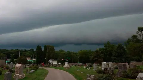  A person walks through Rose Hill Cemetery as a shelf cloud approaches behind them ahead of a severe thunderstorm. The clouds are grey and heavy in the sky. 