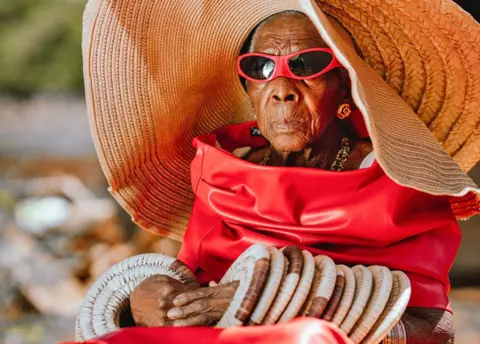 Luxury Media Zambia Margret Chola looks serious as she wears a giant straw hat with straw bangles on both of her arms. He wore red sunglasses and what looked like a red leather jacket