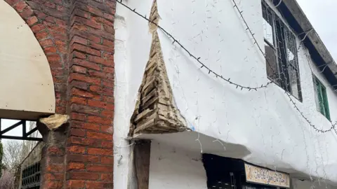 The exterior of a damaged building. A section of red brickwork, arranged in a traditional staggered pattern, is clearly visible. There is a stone block embedded partly in the brick column, protruding slightly. Next to it appears to be a wooden panel or board placed over an opening or archway.