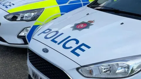 BBC The bonnets of two police cars. The cars are white with blue writing spelling police alongside the Isle of Man Constabulary crest, which is blue and red and features the three legs of Mann. The side panels of one car, which are blue and bright yellow, can be seen.