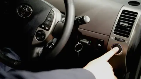 A person sat in the driver's seat of a car pressing a button to start the engine