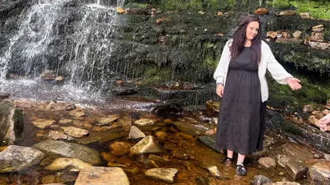 Joshua Wooldridge Bethany Wooldridge in the outdoors. She is stood next to a waterfall and has one hand outstretched. She is wearing a long black dress and a cream cardigan, and has long brown hair. 