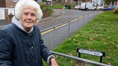 British Heart Foundation Joan stands on the street in front of a metal handrail and a sign which reads "Willett Way". She is looking at the camera and smiling, and is wearing a dark blue coat with the cuffs turned slightly up.