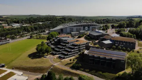 University of Brighton An aerial view of the university's Falmer campus which shows a modern building in the background with a white metal arch reaching over the top of it and in the foreground stepped buildings and brick buildings with solar panels on the roof. There are also sports pitches in the picture and green hills in the background.