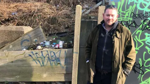 Man standing next to wall with graffiti with beers cans and other rubbish behind broken down fencing 