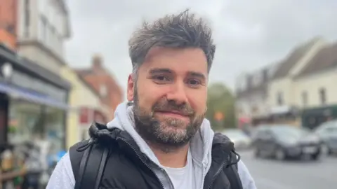 Louise Graham/BBC A man smiles at the camera. He has short gray hair with a gray beard. He wears a black padded vest over a gray hoodie and white t-shirt. He stands outside with shops and cars behind him.