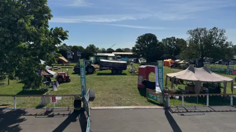 A field includes stalls and a tractor. There are two green banners either side of the entrance to the field near the bottom of the image. There are trees on the left and in the background.