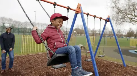 A boy is on a swing and being pushed by a man behind him in a park. It is an autumnal scene. The boy wears a magenta baseball cap and magenta padded jacket with white piping and blue jeans.