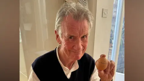 Simon Balch Sir Michael Palin sits at a kitchen table and holds an egg in one hand that he has signed with a pen. He has short grey hair and wears a white shirt with a black vest over the top.