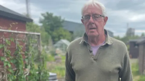 David has grey hair, is wearing a green jumper and is pushing a wheelbarrow filled with woodchopping's through his allotment. 