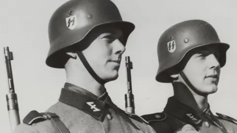 Two members of the Dutch Volunteer Legion of the SS stand side by side, wearing Nazi uniforms, helmets, and with rifles hanging on their shoulders.