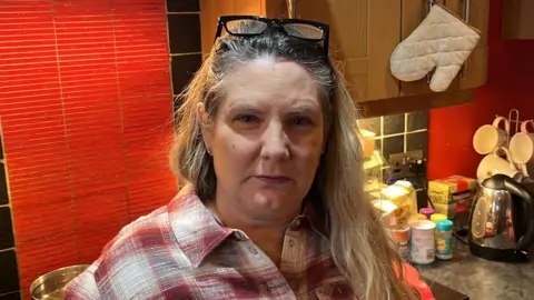 Phil Harrison/BBC A blonde woman standing in a kitchen. She has a button-up shirt on and black glasses on her head.