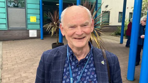 BBC/RICHARD EDWARDS This image shows Green Party councillor Andy Brown smiling straight at the camera. He is standing outside North Yorkshire Council's offices in Selby, wearing a blue patterned shirt, and a blue checked jacket with a metal badge of a bumble bee on its collar.