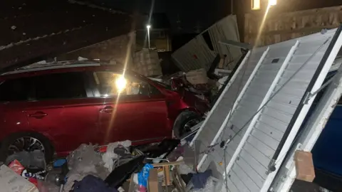 Contributed A red car that has crashed through a garage. The tiled roof of the building is on top of the car and a battered steel garage door is off its hinges. There are boxes and other items that were inside the garage around the wheels of the car.