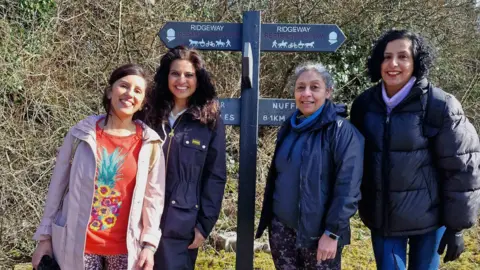 Geeta Ludhra Geeta pictured with three other femal walkers, standing either side of a footpath sign saying "Ridgeway" on the top two arrows (pointing left and right). Other arrows below are concealed. There is a thick hedgerow of brambles and trees behind them. They wear autumn/winter coats and all smiling at the camera. One woman is older, about 60 plus, while the others appear to be in their 30s-50s.