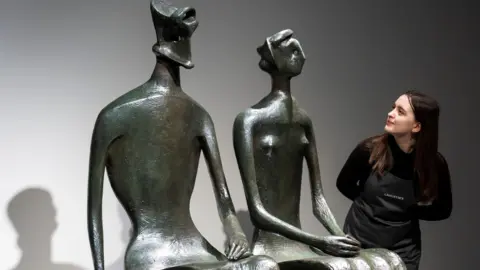 PA Media A view of two bronze figures, with a woman with dark hair, a dark top and a grey apron on peering around one of the figures to look at them both.