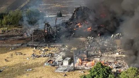 Stephen Huntley/BBC Large plumes of thick black smoke rise into the air from a farm. Orange flames are seen in the wreckage of some buildings. There is shrapnel strewn across a field.