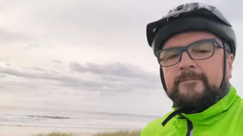 Family A man wearing a cycling helmet, glasses and a light green jacket, takes a selfie. Behind him is a beach, with blades of grass, sand and the sea visible.