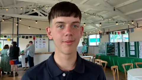 A young man wearing a blue polo shirt smiles. He has short, straight brown hair and blue eyes.