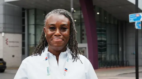 John Fairhall/BBC Ijeoma Okolo wears a white shirt with a lanyard around her neck. She has braided hair and is smiling at the camera. She stands in front of the Gateway building at the University of Essex's Southend campus.