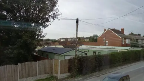 A group of green caravans behind a fence in a residential area with houses behind and a road in front.