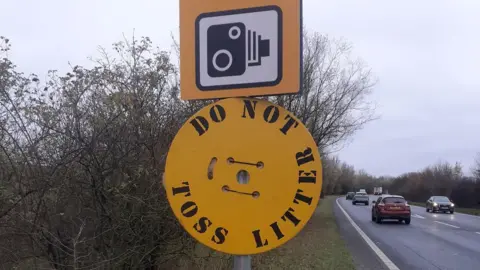 A yellow circle-shaped metal sign with ' do not toss litter' written on it in black, placed on a lamppost on the side of the road.