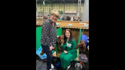 Laura Crombie Two women at Crufts. The one on the left has blonde hair and is wearing a floral shirt and jeans and is smiling at the camera. The second woman is sat down next to a dog in a booth, wearing a green suit.