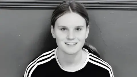 Black-and-white picture of Holly. She is sitting with her back against a wall and is smiling at the camera. Her long hair is in a ponytail and she is wearing a black top with white stripes down the shoulders.