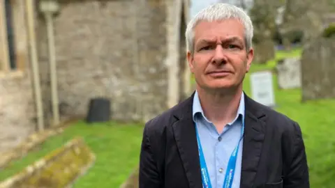 BBC/Seb Cheer A man wearing a light blue shirt and black blazer looks at the camera. He is standing outside a church and has a lanyard on which reads "Healthwatch".