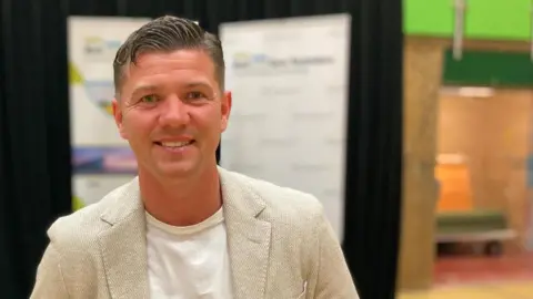Luke Campbell smiles into the camera after winning the mayoral election in Hull and East Yorkshire. He is wearing a beige blazer with a white t-shirt. He is standing in front of a stage that includes a podium and a black background.