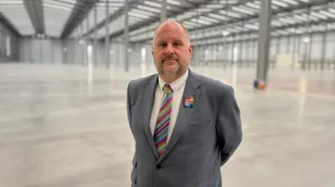 Swindon Borough Council Man in colourful tie, white shirt and grey jacket stood in an empty warehouse.