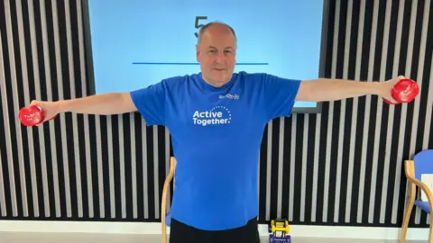 BBC/Richard Edwards Les Wallis, a patient, holds out his arms with weights in his hands