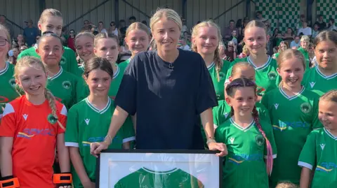 Laura Foster/BBC Leah Williamson sostiene una camiseta enmarcada mientras posa con un equipo de chicas jóvenes