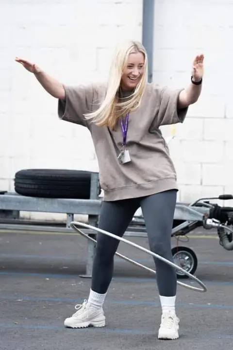 Emily Cooke Photo shows Emily Cooke hula hooping in a playground. She has long blonde hair and is wearing a grey top, leggings and white trainers.