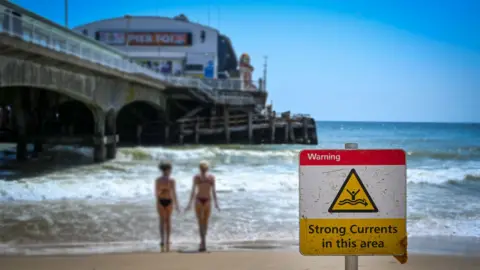 Getty Images A sign warning of strong currents is seen at Bournemouth Pier. Two younf women are at the eadge of the sea, the pier is in the background and on the sand is a red sign that says "Strong currents in this area"