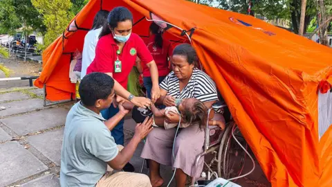 Getty Images A woman sitting in a wheelchair outside a bright orange tent holds a small infant in her arms, an oxygen mask over its face, as a man and a woman tend to the child
