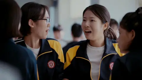 Four high school girls sit together in a cafeteria. They are wearing matching black-and-yellow athletic-school school jackets. Joo-in's friend, left, is turned slightly to the side wearing glasses, while Joo-in faces forward with her mouth open mid-expression 
