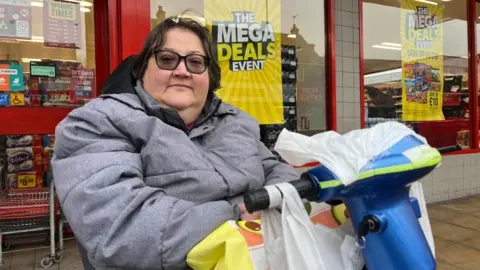 Andrew Turner/BBC Sally Phipps is sitting in her mobility scooter outside Iceland, with bargains advertised in the windows. She has dark hair, has tinted spectacles and is wearing a grey jacket. She has a variety of carrier bags hanging from the handlebars of her scooter