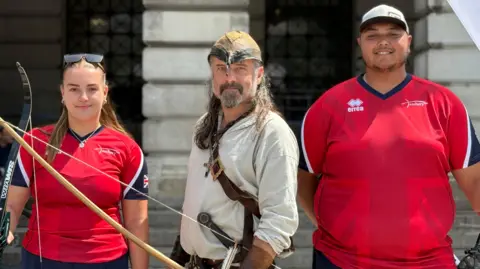 A man and woman in red Team GB archery sports tops stand either side of a man dressed as Robin Hood. He is wearing a cream/white coloured top and is holding a bow and arrow