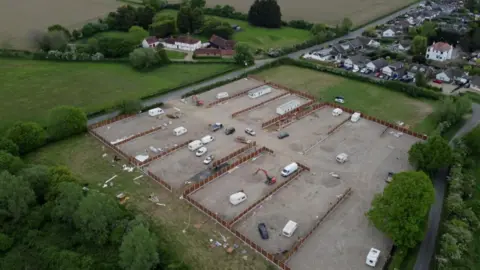 An aerial photo of a traveller site development in Willows Green. A field is mostly covered in hardcore and fencing, with several vehicles on site. There are green fields surrounding the site and houses in the distance