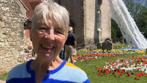 An older woman is smiling to camera. Behind her is an ancient church with the art installation she designed. It shows a waterfall made from old plastic bags flowing from the church tower into a carpet of brightly coloured knitted flowers. The woman has short white hair and is wearing a blue striped cardigan. It is a bright and sunny day.
