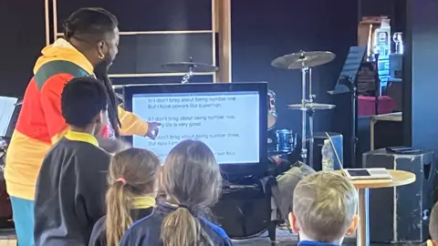 A group of children standing looking at a TV screen, a man is pointing at the screen