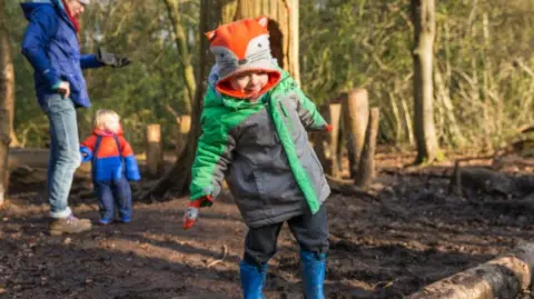 A little boy is balancing on a log on the muddy ground. He is wearing a woolly hat that looks like a fox's face and has on a black and green anorak, trousers and blue boots. Behind him is a woman and a younger child.