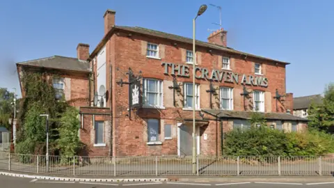 Google A red brick building with the words The Craven Arms at the top. Trees surround the building, alongside some street fences