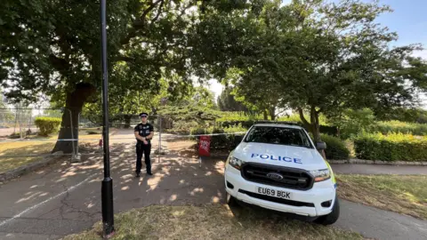 Stuart Woodward/BBC A male police officer standing by a cordon that has been put up in the park.
There is a police car parked nearby too.