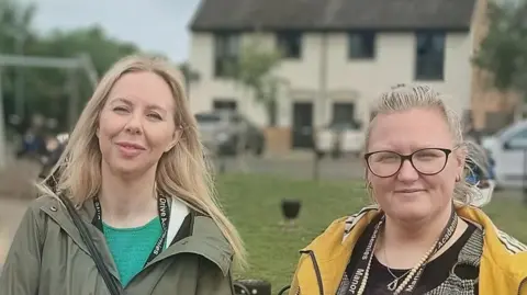 Laura Owen and Fern Rowe looking at the camera, while in a small playground on the Manor Drive estate. Laura has long blonde hair and is wearing a dark green coat over a turquoise blouse. Fern also has long blonde hair and is wearing glasses, and a yellow coat over a black top.