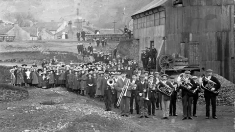 Beamish, The Living Museum of the North A black and white image of about 100 people standing in front of a tall Mill building and looking at the camera. Most of them are wearing hats and long coats. About a dozen men at the front are carrying brass instruments. There are houses in the background.