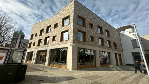 Aimee Dexter/BBC A large three-storey building is in the middle. The first floor has large windows and an entrance on the left. The top two floors have a diamond pattern on the outside. 