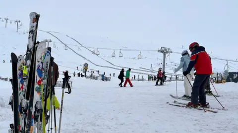 Skis stand upright in the snow beside a busy ski slope, where skiers prepare to set off near chairlifts and lift towers under overcast winter skies.